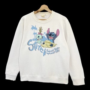 K&ouml;nnte beinhalten: Wei&szlig;er Pullover mit einem blauen und rosa Stitch- und Angel-Motiv. Der Text "Stitch Tropical Trouble Experiment 626" ist unter dem Motiv gedruckt.