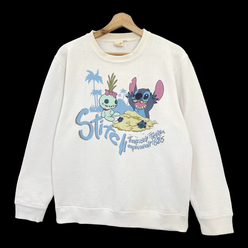 K&ouml;nnte beinhalten: Wei&szlig;er Pullover mit einem blauen und rosa Stitch- und Angel-Motiv. Der Text "Stitch Tropical Trouble Experiment 626" ist unter dem Motiv gedruckt.
