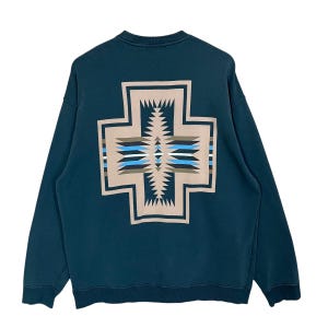 May include: Teal sweatshirt with a large, geometric cross design on the back. The cross features a beige border and intricate patterns in shades of blue, brown, and white. The long-sleeved top is made of a soft material.
