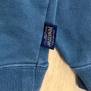 以下が含まれることがあります： リブ編みの裾と袖口が付いた、ティール色のスウェットシャツのクローズアップ。ブランド名「Pendleton」のネイビーブルーのタグが衣服に縫い付けられています。タグには「Warranted to be a...」と「Pendleton Woolen Mills Since 1863」の文字も含まれています。