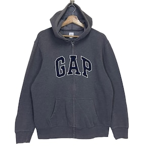 Könnte beinhalten: Ein anthrazitfarbener Kapuzenpullover mit Reißverschluss und dem Wort "GAP" in marineblauen Blockbuchstaben mit weißer Umrandung auf der Brust. Der Hoodie hat eine Kapuze mit Kordelzug, einen durchgehenden Reißverschluss und zwei Vordertaschen. Er wird auf einem Holzkleiderbügel präsentiert.
