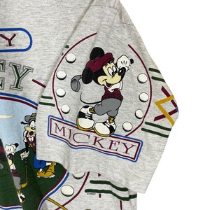 Puede incluir: Camiseta gris claro con un dise&ntilde;o de golf de Mickey Mouse. La camiseta presenta a Mickey Mouse con un atuendo de golf, rodeado de pelotas de golf y la palabra "MICKEY". La palabra "GOLF" tambi&eacute;n se muestra en un dise&ntilde;o azul y verde.