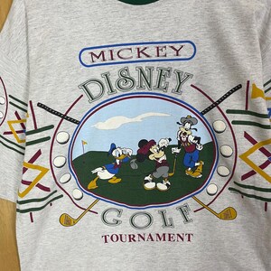 Puede incluir: Camiseta gris con un gr&aacute;fico colorido de Mickey Mouse, el Pato Donald y Goofy jugando al golf. La camiseta tiene las palabras "Mickey Disney Golf Tournament" impresas. La camiseta tiene un cuello verde y dise&ntilde;os geom&eacute;tricos coloridos.