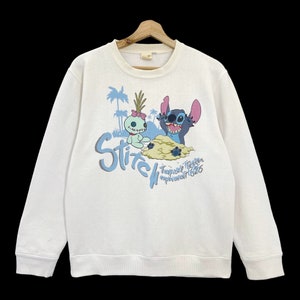 K&ouml;nnte beinhalten: Wei&szlig;er Pullover mit einem blauen und gr&uuml;nen Motiv von Stitch und Angel. Der Text "Stitch" ist in Blau und der Text "Tropical Tracker Experiment 626" ist in Blau und Schwarz.