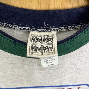 Puede incluir: Primer plano del cuello de una camiseta gris con ribete azul marino y verde. Una etiqueta blanca con texto negro y un logotipo est&aacute; adjunta. La etiqueta dice "MADE IN U.S.A." y "Large".
