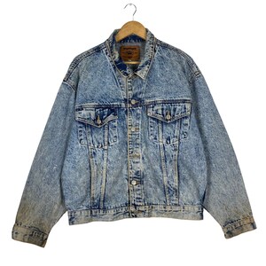May include: A light blue acid wash denim jacket with a classic collar, button-down front, and multiple pockets. The jacket is hanging on a wooden hanger. The denim has a faded, worn appearance.