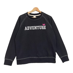 May include: Black crew neck sweatshirt with the word "ADVENTURE" in white block letters. A pink heart-shaped logo is above the word. White stitching accents the shoulders and neckline. The sweatshirt is hanging on a wooden hanger.