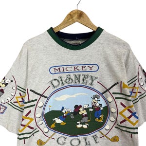 Puede incluir: Camiseta gris claro con un dise&ntilde;o de Mickey Mouse Disney Golf. El gr&aacute;fico incluye a Mickey, Donald y Goofy jugando al golf. La camiseta tiene un cuello verde y azul marino y el texto "Mickey Disney Golf".