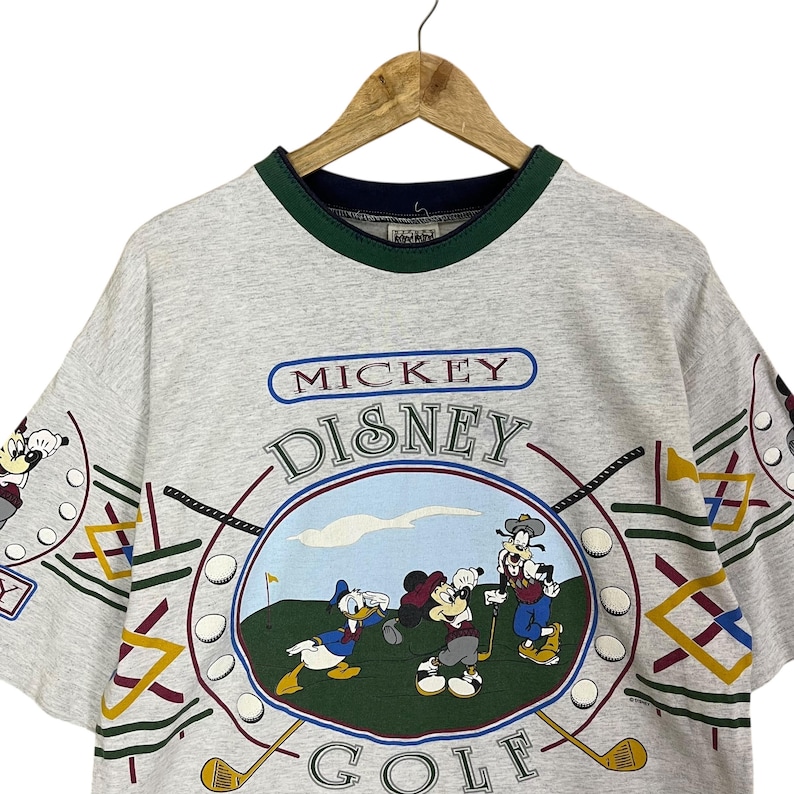 Puede incluir: Camiseta gris claro con un dise&ntilde;o de Mickey Mouse Disney Golf. El gr&aacute;fico incluye a Mickey, Donald y Goofy jugando al golf. La camiseta tiene un cuello verde y azul marino y el texto "Mickey Disney Golf".