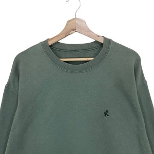 Pu&ograve; includere: Una felpa verde salvia con girocollo e maniche lunghe. La felpa presenta un piccolo logo nero ricamato sul petto sinistro. &Egrave; appesa a una gruccia di legno su sfondo bianco.