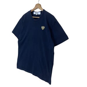 May include: Navy blue t-shirt with a small embroidered heart-shaped logo featuring eyes. The shirt has short sleeves and a crew neck. The brand name "PLAY" is visible on a tag at the neckline.