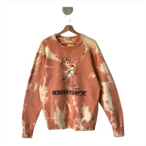 May include: A tie-dye sweatshirt in shades of orange and cream, featuring a graphic of Snoopy in a pilot's outfit. The word "Snoopy" is printed in black block letters below the character. The sweatshirt is hanging on a wooden hanger.