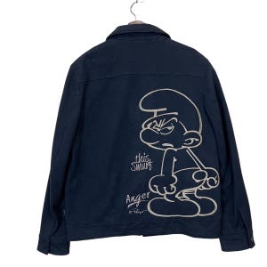 May include: Navy blue denim jacket featuring a white outline illustration of an angry Smurf character on the back. The text "this Smurf" and "Anger" are also visible. The jacket has a button-up front and two chest pockets.