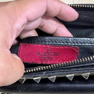 May include: Close-up of a black leather handbag with silver-toned hardware. A red tag with the brand name "Valentino" is visible. The bag has a zipper closure and decorative studs.
