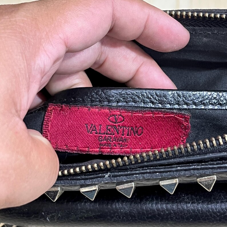 May include: Close-up of a black leather handbag with silver-toned hardware. A red tag with the brand name "Valentino" is visible. The bag has a zipper closure and decorative studs.