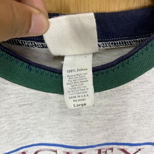 Puede incluir: Primer plano de un cuello de camiseta gris con una etiqueta blanca. La etiqueta dice "100% Algod&oacute;n" e incluye instrucciones de lavado. La camiseta tiene un ribete azul marino y verde y est&aacute; etiquetada como "Large".