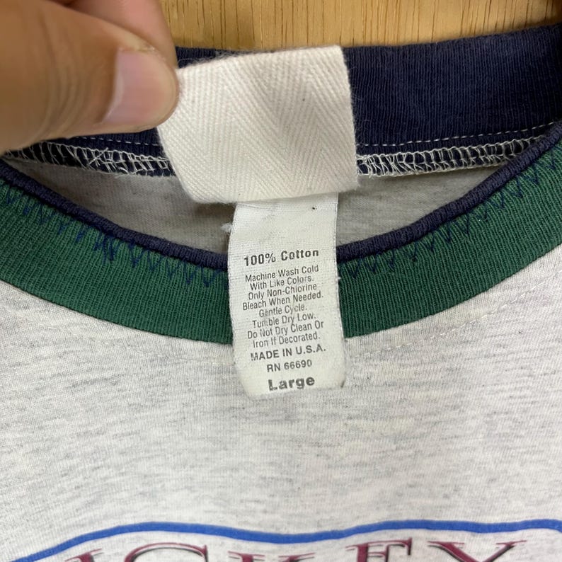 Puede incluir: Primer plano de un cuello de camiseta gris con una etiqueta blanca. La etiqueta dice "100% Algod&oacute;n" e incluye instrucciones de lavado. La camiseta tiene un ribete azul marino y verde y est&aacute; etiquetada como "Large".