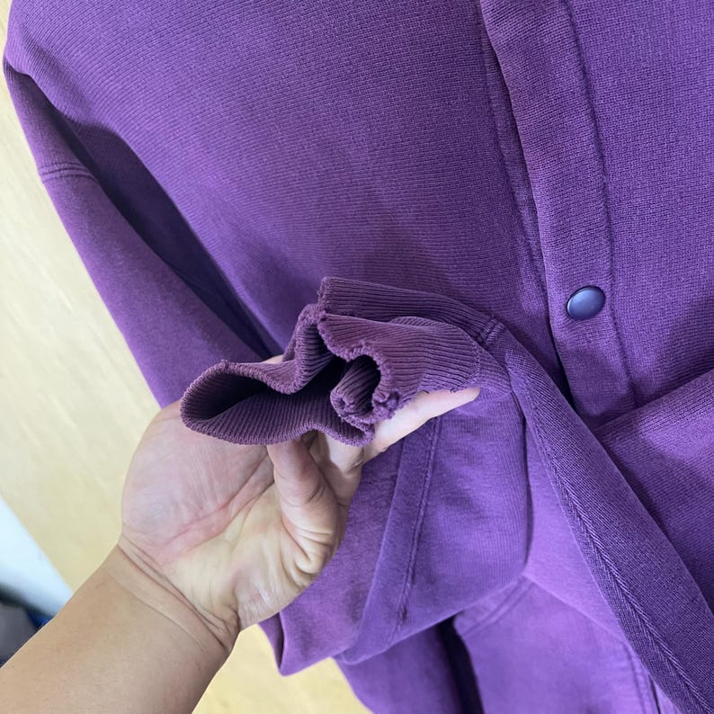 May include: A close-up of a purple sweatshirt with a ribbed cuff. The sweatshirt has a button closure and a soft, textured appearance. The fabric is a solid, deep purple color.