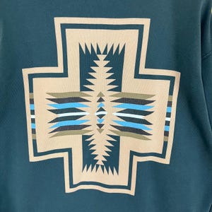 以下が含まれることがあります： ベージュ、青、黒の大きな幾何学模様が特徴のティールスウェットシャツ。 デザインは、複雑なパターンと線を持つ十字架の形をしています。 スウェットシャツは柔らかい素材で作られています。