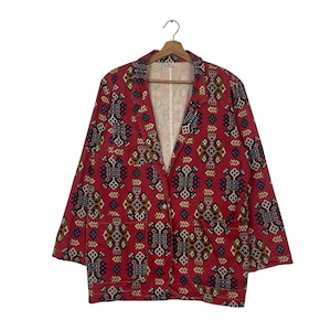 May include: A red blazer with a geometric pattern in shades of blue, yellow, and brown. The blazer has a notched collar, two front pockets, and long sleeves. It is hanging on a wooden hanger.