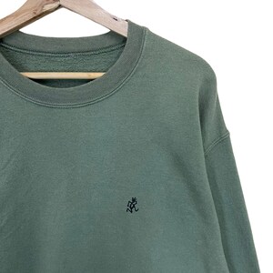 Pu&ograve; includere: Una felpa verde salvia con girocollo e maniche lunghe. Un piccolo logo nero ricamato &egrave; sul petto sinistro. La felpa &egrave; appesa a una gruccia di legno su sfondo bianco.