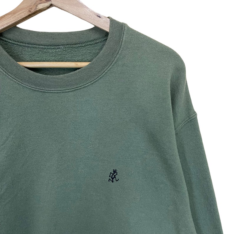 Pu&ograve; includere: Una felpa verde salvia con girocollo e maniche lunghe. Un piccolo logo nero ricamato &egrave; sul petto sinistro. La felpa &egrave; appesa a una gruccia di legno su sfondo bianco.