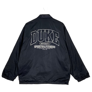 Może przedstawiać: Czarna nylonowa kurtka z napisem "DUKE" dużymi, białymi literami na plecach. Poniżej napis "UNIVERSITY SPORTS & FITNESS" oraz słowa "SINCE 1839". Kurtka ma kołnierz.