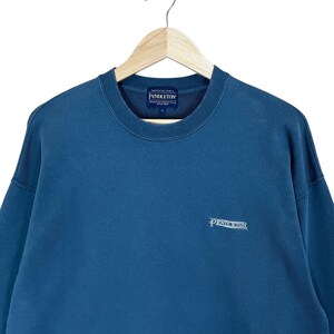 以下が含まれることがあります： 胸と右下に白で「Pendleton」のブランド名が刺繍された、青色のクルーネックのスウェットシャツ。白い背景に、木製のハンガーにスウェットシャツが掛けられています。