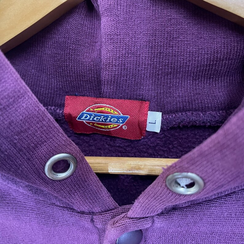 May include: Close-up of a purple Dickies hoodie. The hoodie features a red tag with the Dickies logo and a size "L" label. The fabric is a solid purple color, and the hoodie has metal eyelets and a button closure.