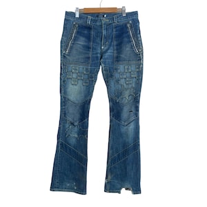 May include: Blue denim jeans with distinctive patchwork and star-shaped details. They have front pockets, side zippers, and a flared leg. The jeans are displayed on a wooden hanger.