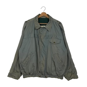 Könnte beinhalten: Hellgraue Bomberjacke mit Kragen und zwei Vordertaschen. Die Jacke hat Knopfverschlüsse an den Schultern und ein grünes Futter. Die Jacke hängt an einem Holzkleiderbügel.