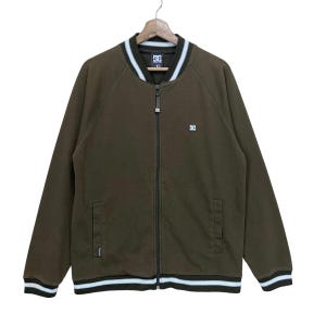 May include: A brown zip-up jacket with white and black striped trim on the collar, cuffs, and hem. The jacket features a small logo on the chest and a full-length zipper. The jacket is hanging on a wooden hanger.