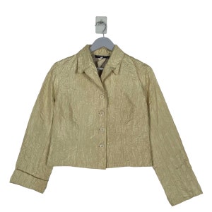 May include: A light beige jacket with a textured surface. The jacket has a collared neckline, long sleeves with folded cuffs, and a button-down front. The jacket is hanging on a gray hanger.