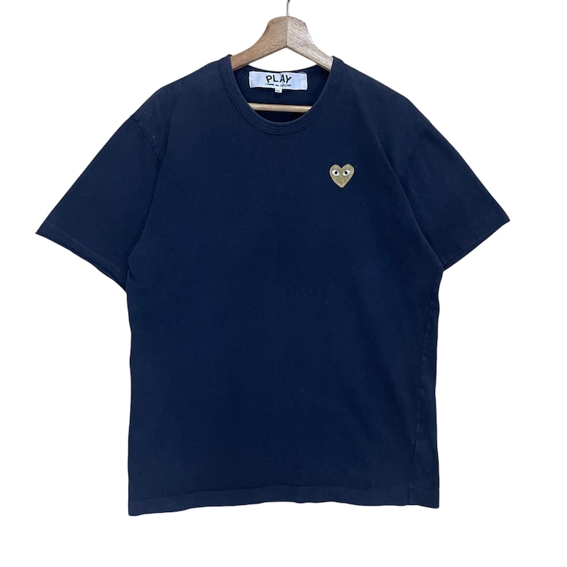 May include: Navy blue t-shirt with a small, embroidered gold heart with eyes on the left chest. The shirt has a crew neck and short sleeves. The word "PLAY" is printed on a white tag at the neckline.