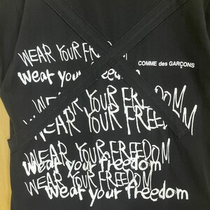 May include: Black t-shirt with white text that says "Wear Your Freedom" repeated multiple times. The text is written in a graffiti style. The shirt has two black straps that cross over the front. The text "Comme des Gar&ccedil;ons" is visible in the top right corner.