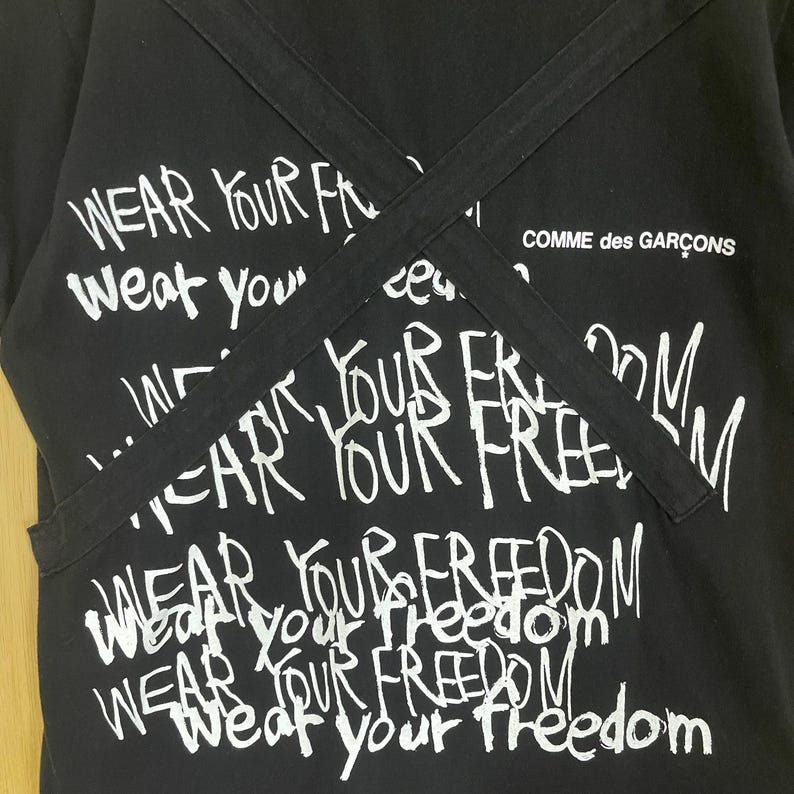 May include: Black t-shirt with white text that says "Wear Your Freedom" repeated multiple times. The text is written in a graffiti style. The shirt has two black straps that cross over the front. The text "Comme des Gar&ccedil;ons" is visible in the top right corner.