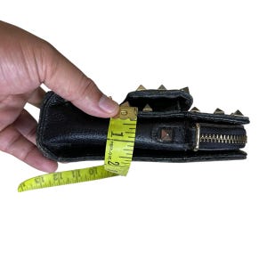 May include: Black leather wallet with gold-colored pyramid studs and a zipper closure. The wallet is being measured with a yellow measuring tape, showing a width of approximately 1.5 inches.