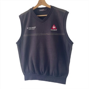 May include: A black sleeveless sweater vest with a white v-neck and a white embroidered line across the chest. The vest has a red rooster logo with the text "le coq sportif" embroidered on the front.