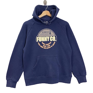 May include: Navy blue hooded sweatshirt with a front pocket and drawstring hood. The sweatshirt features a circular graphic with the text "HAWAII NORTH SHORE FUNNY CO. I WAS BORN AND RAISED IN HAWAII SINCE 1993."