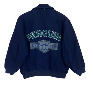 Puede incluir: Una chaqueta bomber azul marino con un logotipo de pingüino verde y blanco en la espalda. El logotipo dice "Penguin" en un arco verde sobre el pingüino y "Since 1886" en un rectángulo blanco debajo del pingüino.