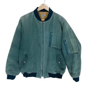 May include: A teal bomber jacket featuring a full-length front zip, a dark navy ribbed collar, cuffs, and hem. The jacket includes two lower snap-button pockets, a zipped chest pocket, and a zipped utility pocket on the left sleeve.