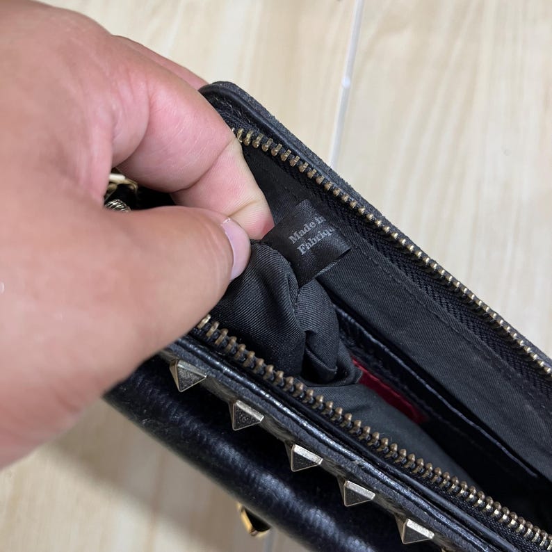 May include: Black leather wallet with gold-colored pyramid studs along the bottom edge. The wallet has a zipper closure and a tag inside that reads "Made in Fabrique".