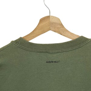Pu&ograve; includere: Felpa verde oliva con scollo tondo, appesa a una gruccia di legno. Il marchio "GRAMICCI" &egrave; stampato in nero sul lato inferiore destro della felpa.