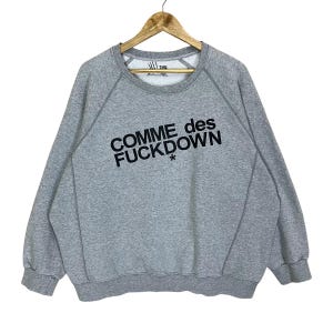 May include: A grey sweatshirt featuring the text "COMME des FUCKDOWN" in black. The sweatshirt has a crew neck and long sleeves. The garment is suspended from a wooden hanger.