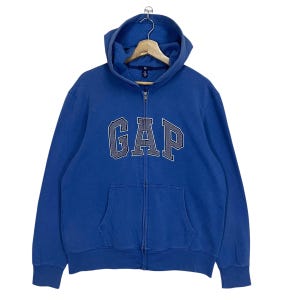 Puede incluir: Una sudadera azul con cremallera y la palabra "GAP" en letras grises en el pecho. La sudadera tiene una capucha con cordón y un bolsillo delantero. La prenda está colgada de una percha de madera.