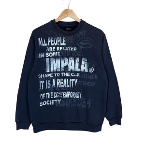May include: A navy blue sweatshirt featuring white text including "IMPALA", "IT IS A REALITY", and "OF THE CONTEMPORARY SOCIETY". The sweatshirt has a crew neck and long sleeves. The text also includes "ALL PEOPLE ARE RELATED OF".