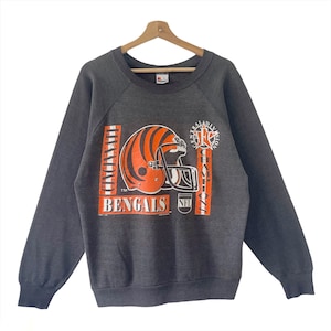 May include: A gray sweatshirt with a vintage Cincinnati Bengals logo. The logo features a football helmet with orange and black stripes, the team name "Bengals" and "Central Division Champions" text. The sweatshirt has a crew neck and long sleeves.