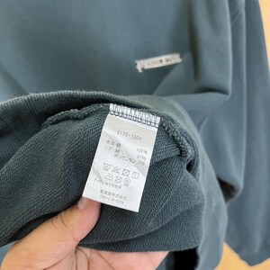以下が含まれることがあります： 襟元近くにタグが見えるティール色のスウェットシャツ。タグには番号「2175-7204」と、生地の組成とケアの手順を示す日本語のテキストが表示されています。スウェットシャツはシンプルなデザインです。