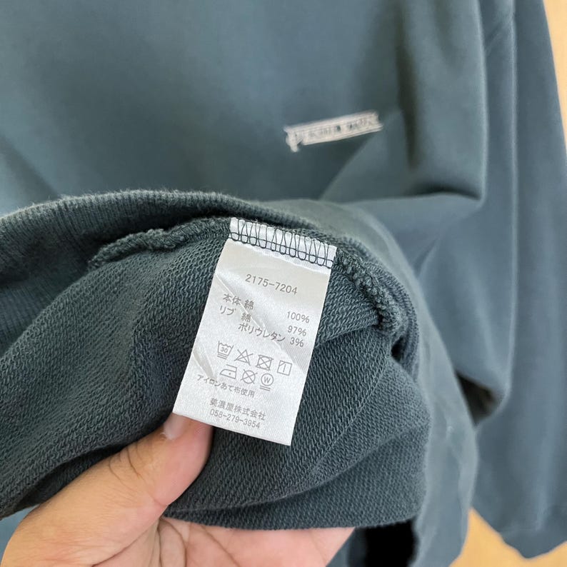 以下が含まれることがあります： 襟元近くにタグが見えるティール色のスウェットシャツ。タグには番号「2175-7204」と、生地の組成とケアの手順を示す日本語のテキストが表示されています。スウェットシャツはシンプルなデザインです。