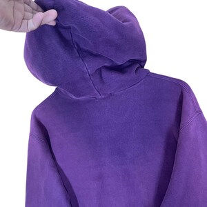 May include: A close-up of a purple hoodie with a hood. The hoodie is made of a soft material and has a drawstring. The image is taken against a white background.